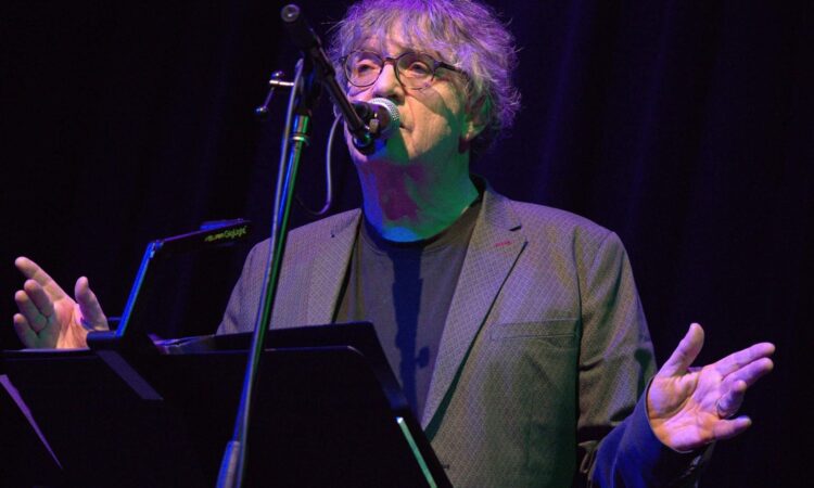The image shows Paul Muldoon reading at a podium
